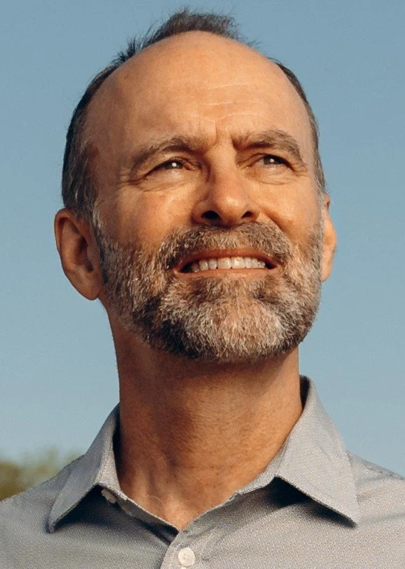 A middle-aged man with a beard and short hair smiling outdoors against a blue sky.