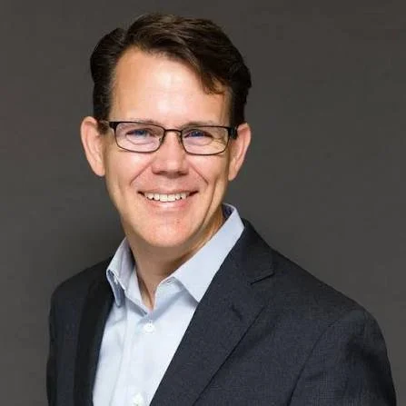 Portrait of a smiling man wearing glasses, a white shirt, and a dark suit against a dark gray background.