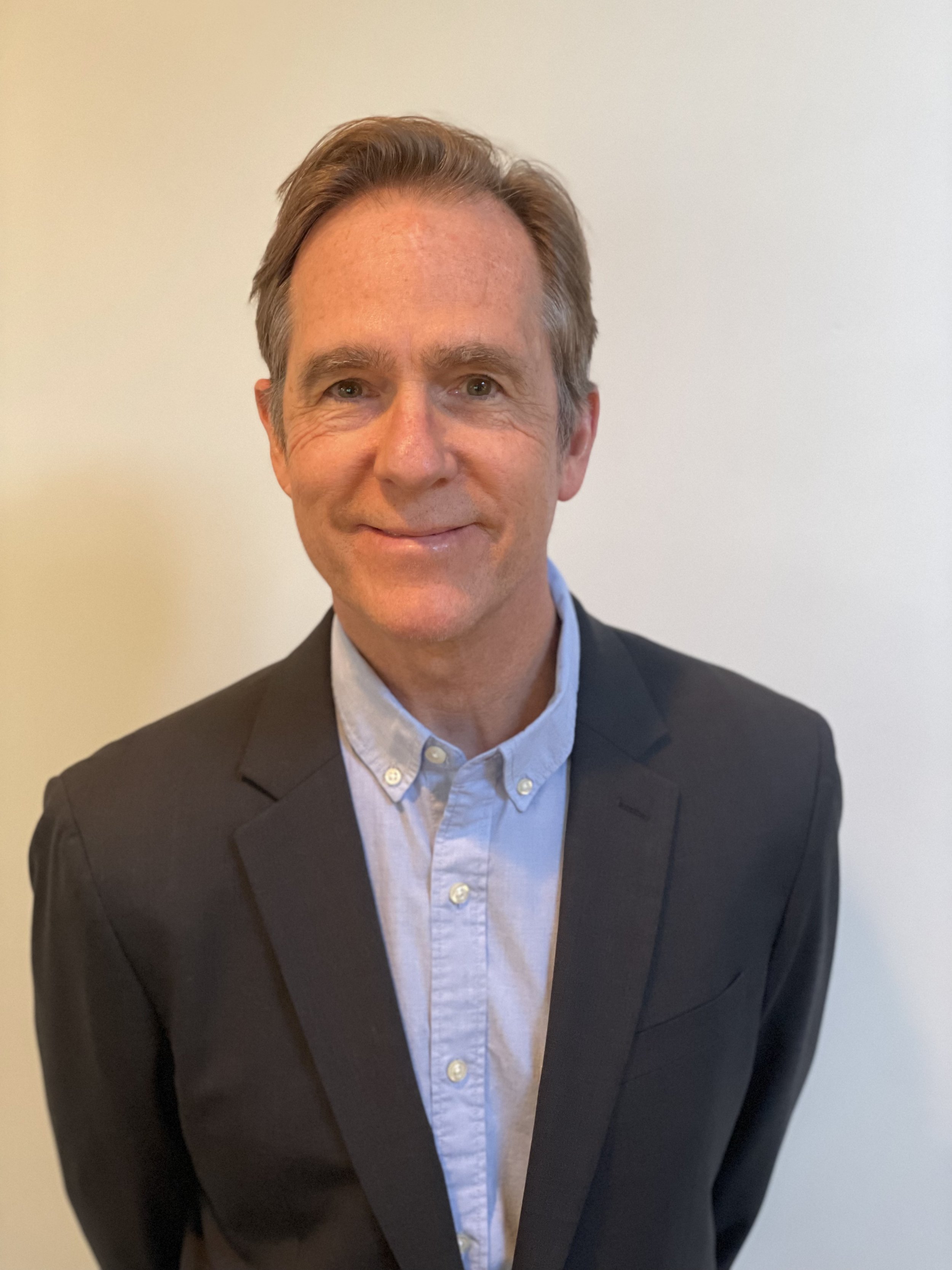 A man with light brown hair, partly gray, wearing a black suit jacket and light blue dress shirt, standing against a plain off-white wall.