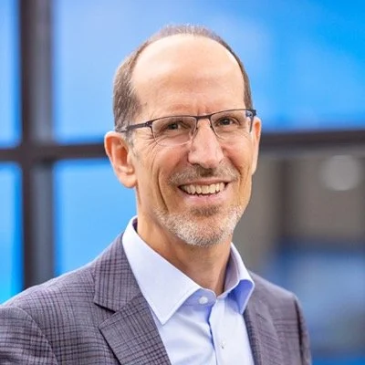 A smiling man in glasses with a beard, wearing a blue shirt and plaid blazer, standing in front of a blue-tinted window.