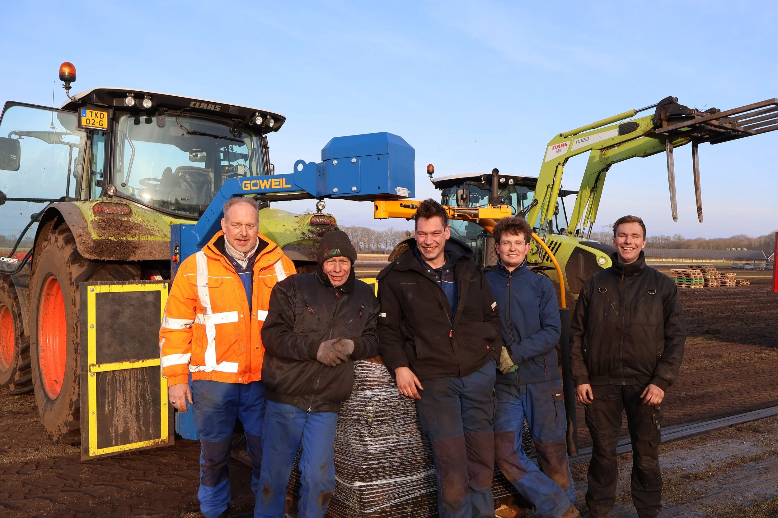 Vijf mannen staan voor landbouwvoertuigen en machines op een veld, met een heldere blauwe hemel op de achtergrond.