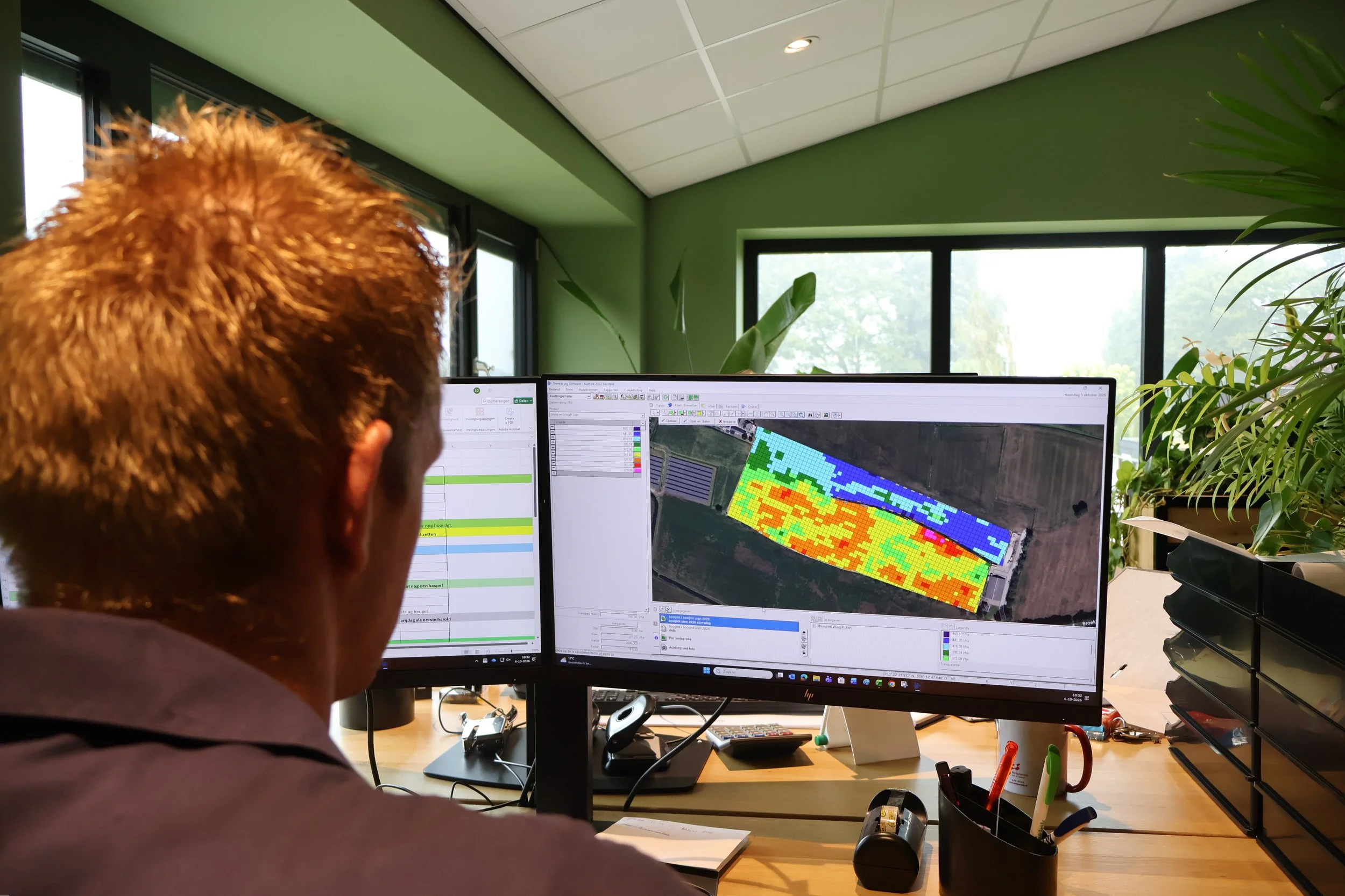 Persoon met rood haar die op kantoor achter een computer werkt, met een monitor die een satellietkaart met kleurrijke gegevens toont, omringd door planten en kantoorbenodigdheden.