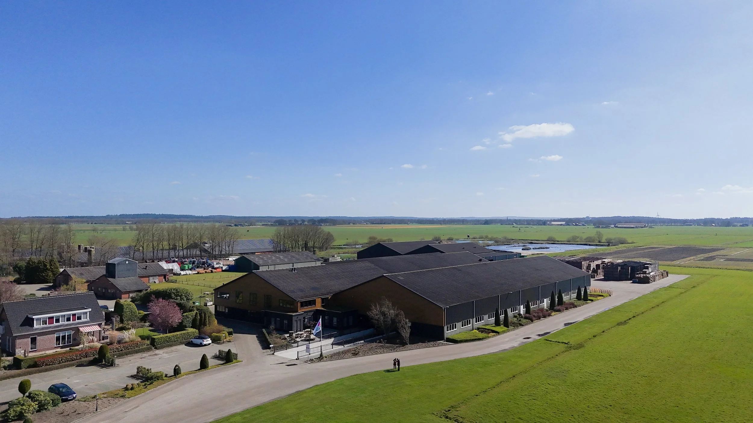 Landschap met woningen, landbouwgebouwen, groene velden, en een open blauwe lucht met enkele kleine wolken