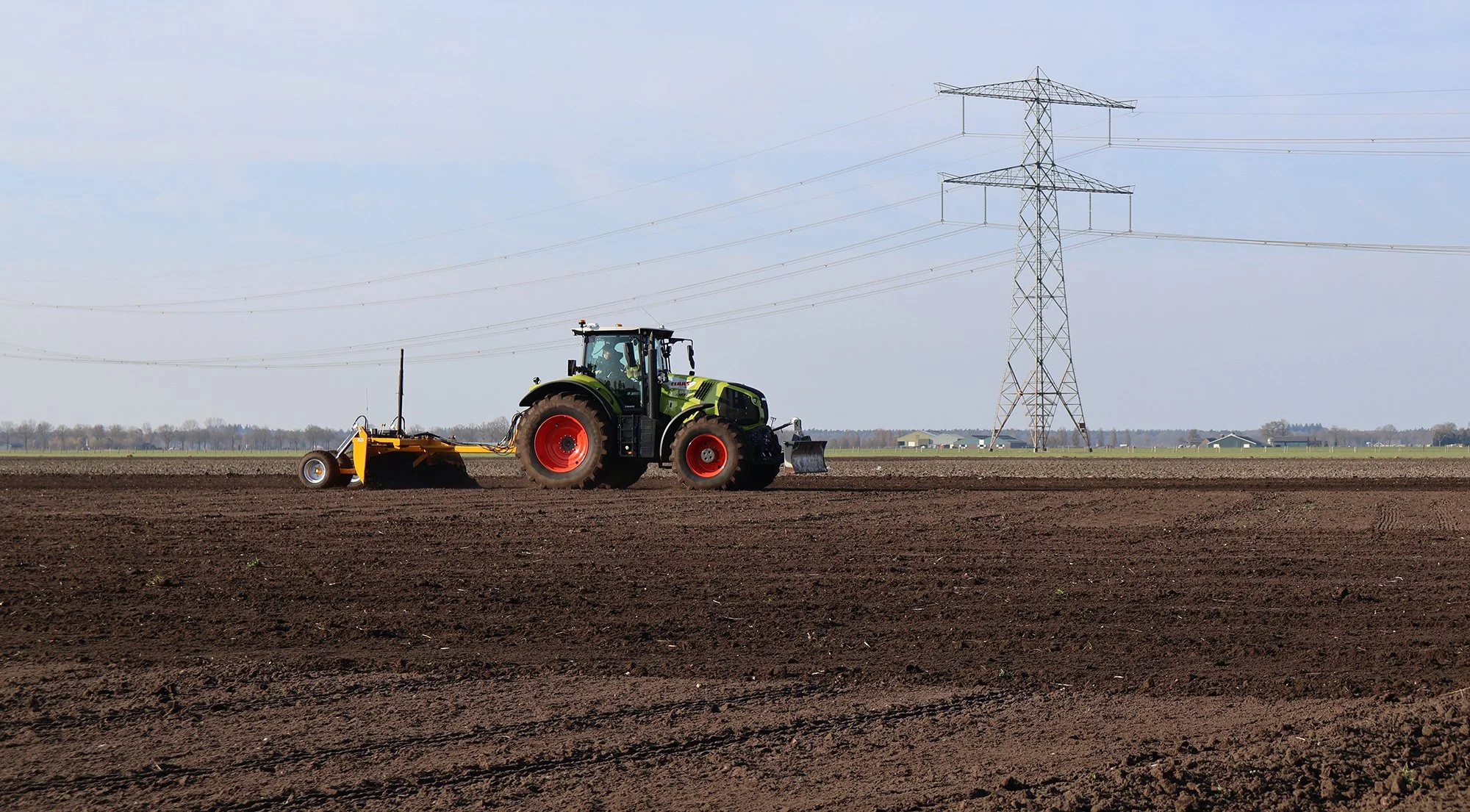 Een tractor werkt op een akker met een grote hoogspanningsmast op de achtergrond.