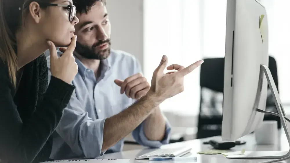 Two colleagues collaborating at a desk, looking at a computer screen; one person is pointing at the screen while the other is thinking with a hand on their chin.
