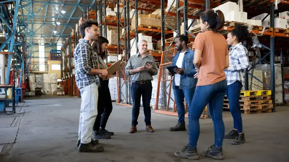 A group of people having a discussion in a warehouse with shelves stacked with boxes and pallets.