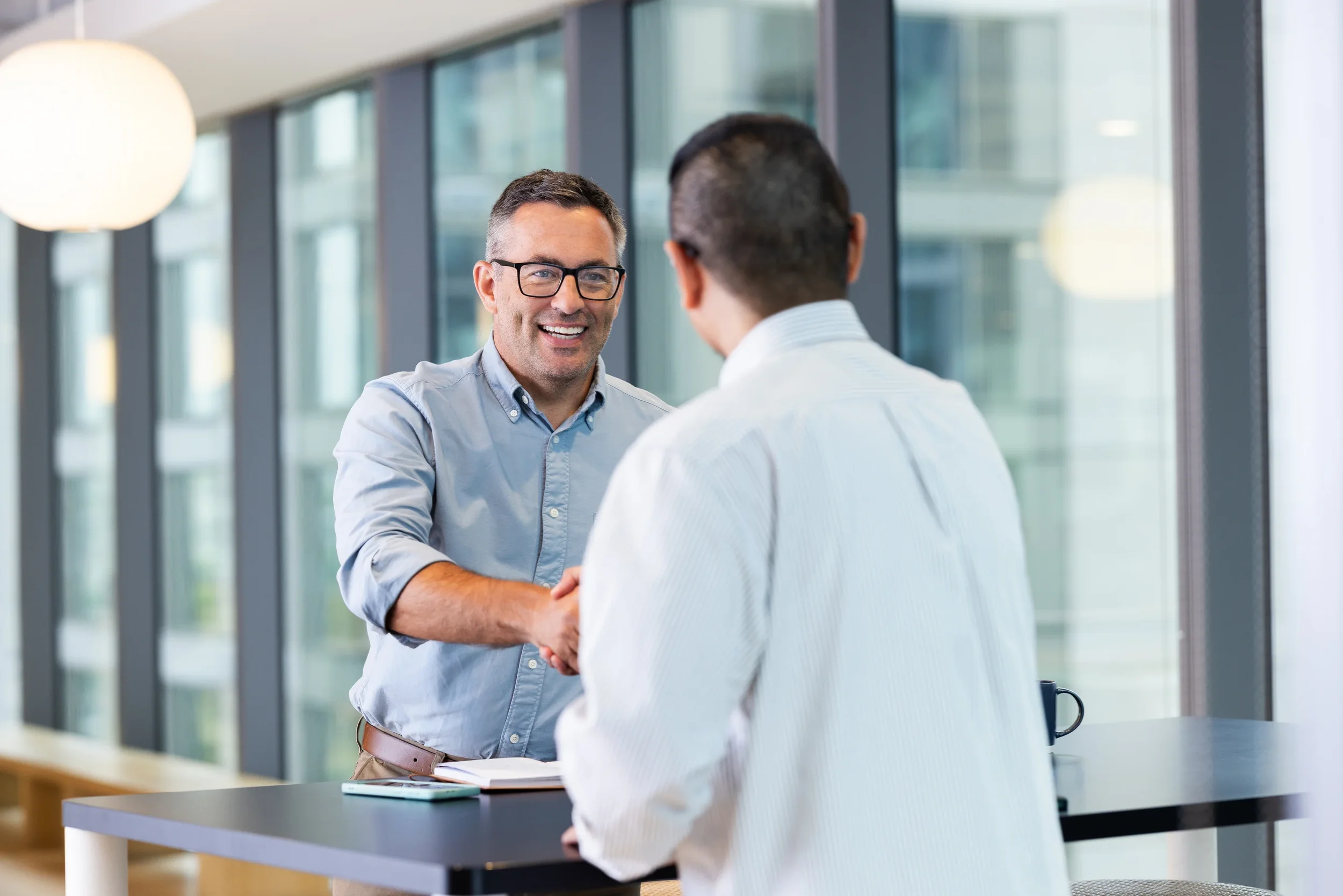 Two men shaking hands in a modern office with large windows, one wearing glasses and a light blue shirt, smiling, the other appears to be in a white shirt, with books and electronic devices on the table.