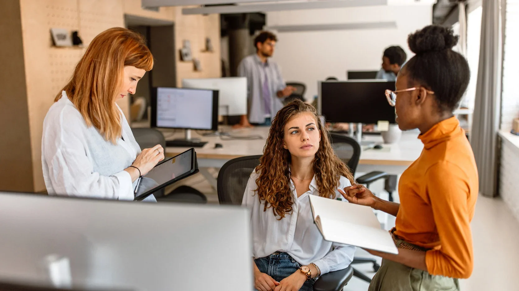 A diverse group of young professionals in an office engaging in a discussion, with a woman sitting and listening, a woman standing and pointing at her, and a man standing in the background using a tablet, surrounded by desks and computers.