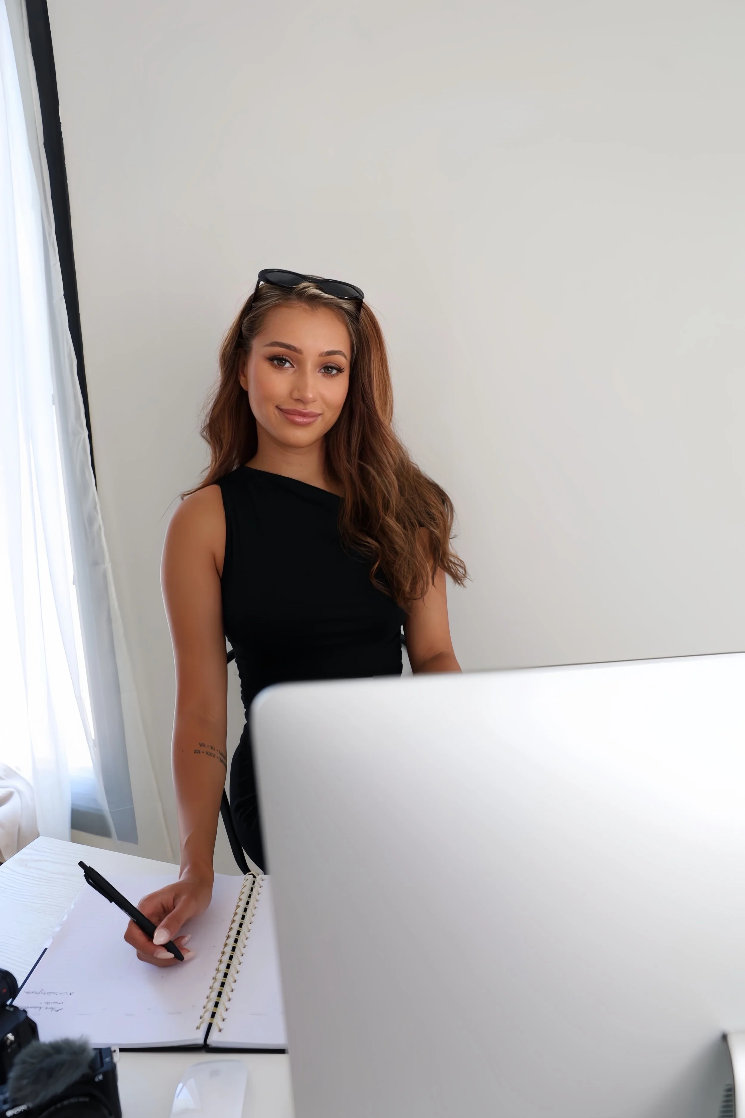 Young woman with long wavy brown hair, wearing a black sleeveless top, sitting at a white desk in a brightly lit room, smiling at the camera with sunglasses resting on her head, taking notes in a notebook.
