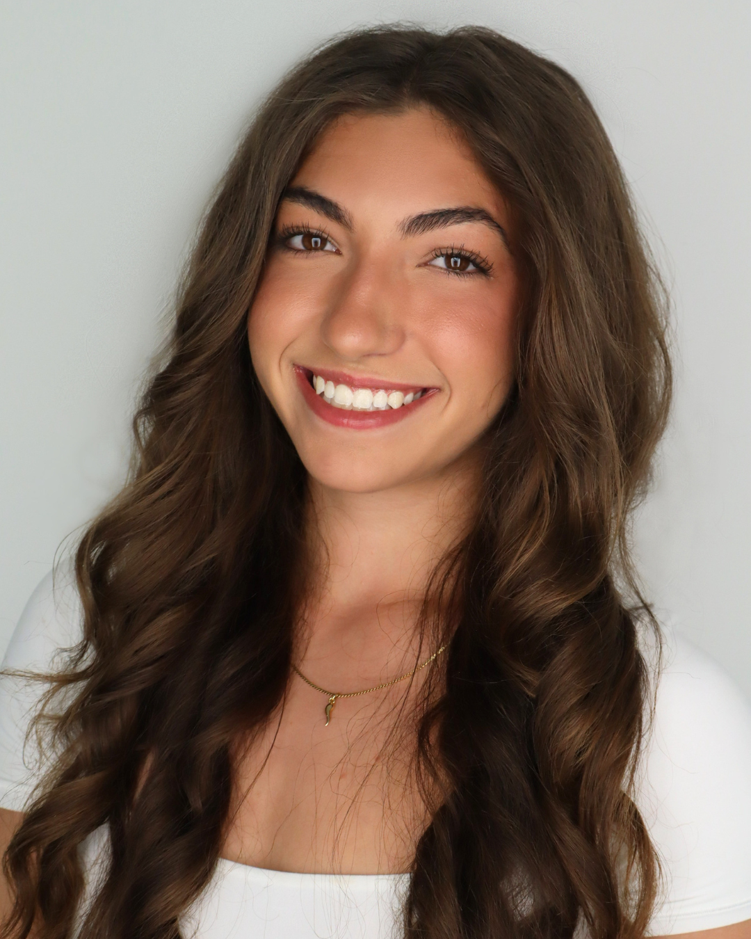 Young woman with long wavy brown hair, smiling, wearing a white top and a gold necklace with a small pendant.