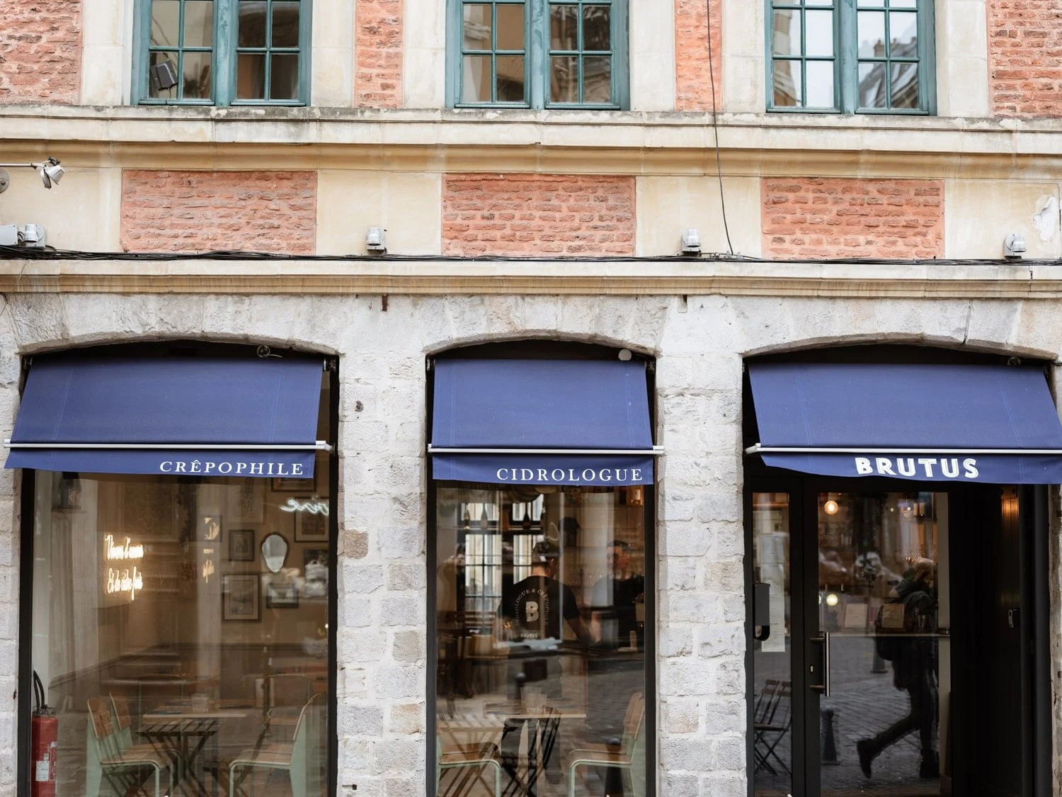 Façade d'un café ou restaurant avec trois grandes fenêtres et panneaux marron foncé. Au-dessus, trois volets verts, et une façade en briques et pierre. Sur les enseignes des vitres, il est écrit "Crépophile", "Cidrologue" et