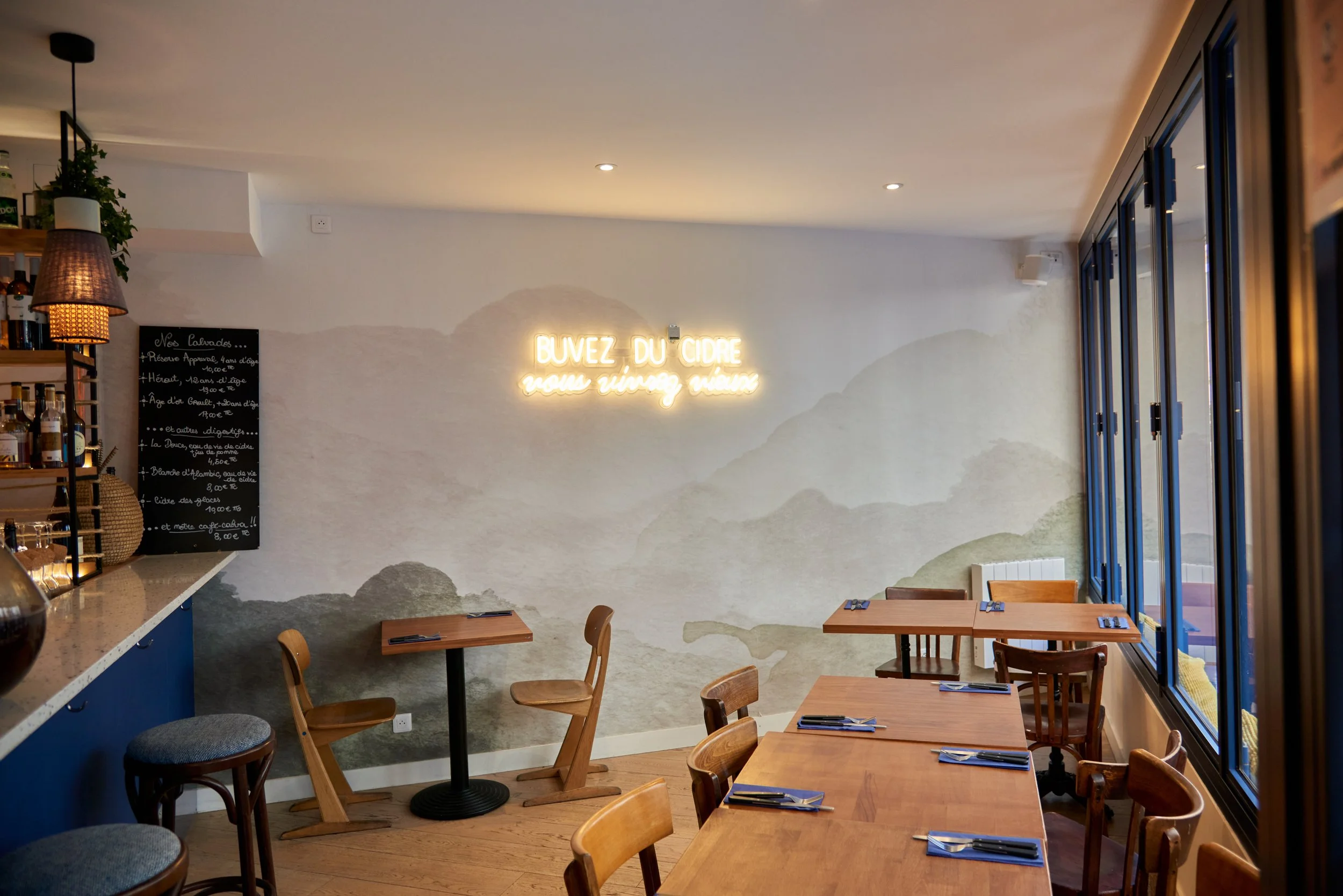 Intérieur convivial d'un restaurant ou café avec tables en bois, chaises, mur avec peinture de montagnes, enseigne lumineuse en français, grande fenêtre laissant entrer la lumière naturelle.