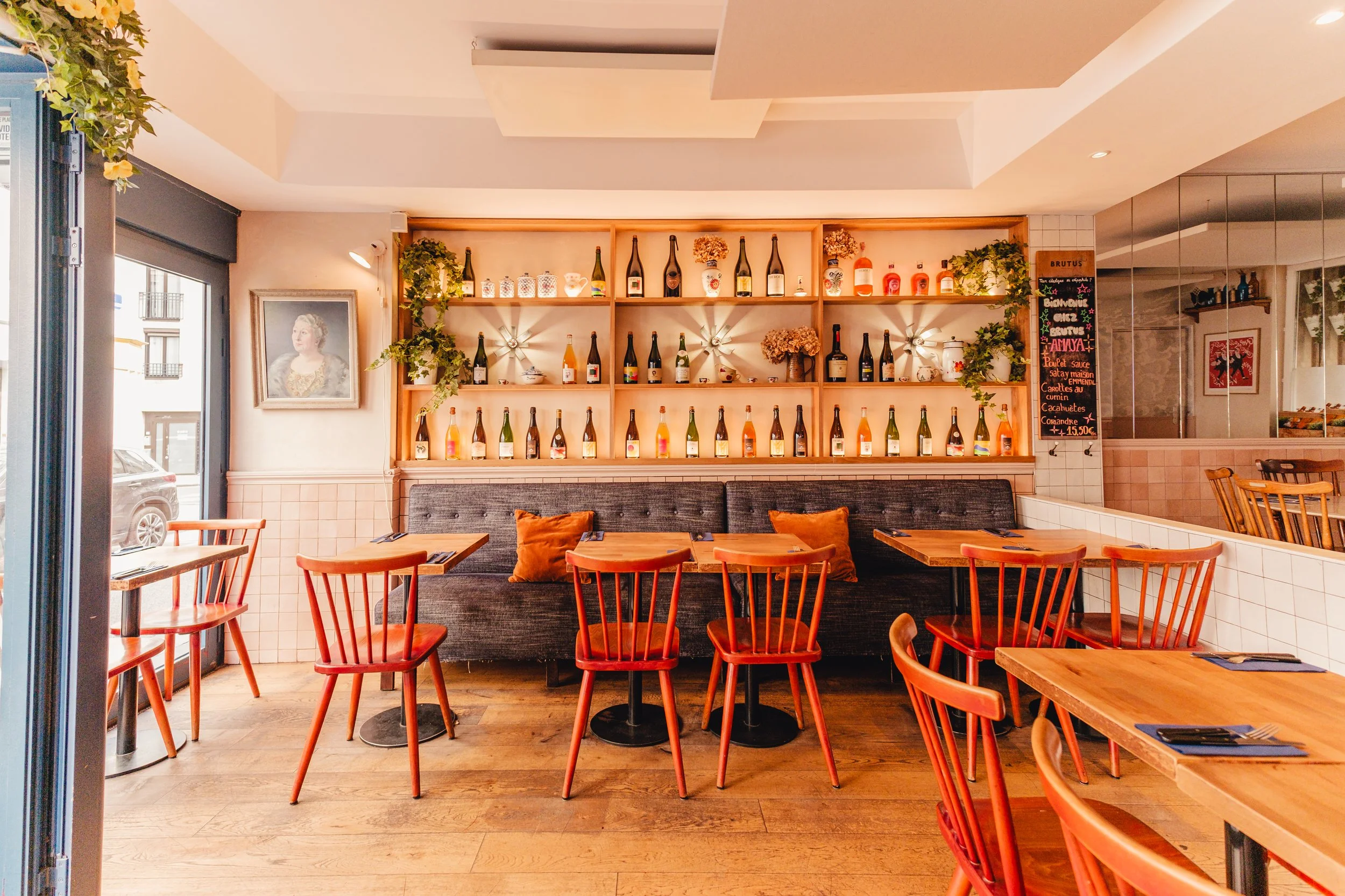 Intérieur d'un restaurant avec des tables en bois, des chaises en bois de couleur rouge, un mur avec étagères en bois contenant des bouteilles de champagne, des vases avec des fleurs séchées, un tableau noir avec un menu, et un cadre avec un portrait de femme. La salle est lumineuse et décorée avec des plantes vertes.