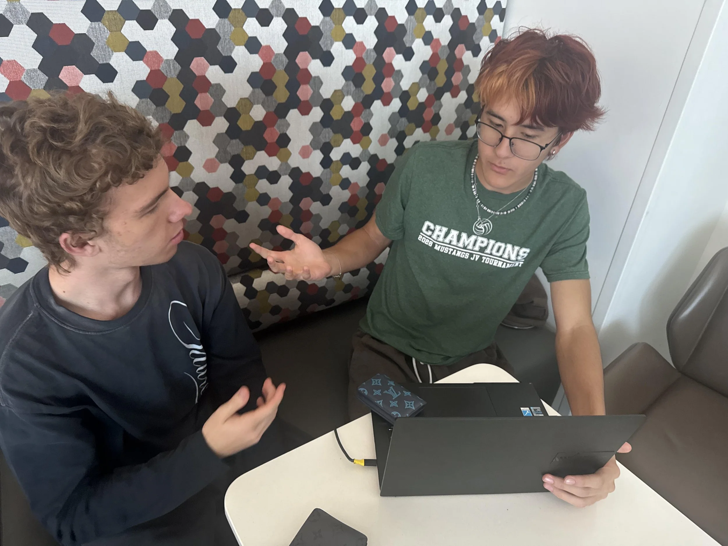 Two young men sit at a table with laptops, one with curly blonde hair and the other with red, wavy hair. The man with red hair is gesturing with his hand, he is wearing glasses, a green T-shirt that says 'Champions 2023 Mustangs JV Tournament,' and a necklace. The background features a patterned wall with hexagon shapes in black, red, gray, and gold.