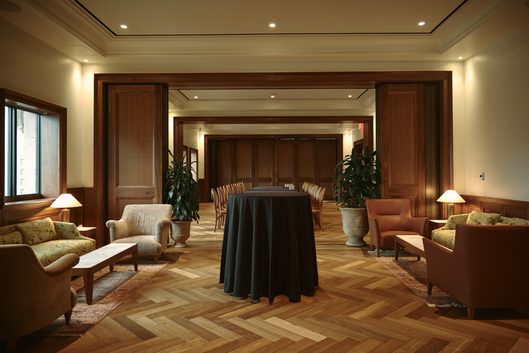 Elegant lounge area with wooden paneling, sofas, armchairs, side tables, lamps, potted plants, and a table with a black tablecloth in the center, featuring a herringbone wood floor and large windows.