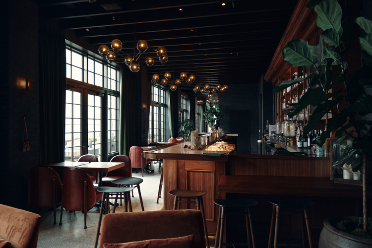 Interior of a stylish bar or restaurant with large windows, modern lighting fixtures, a wooden bar counter, and seating areas with chairs and tables.