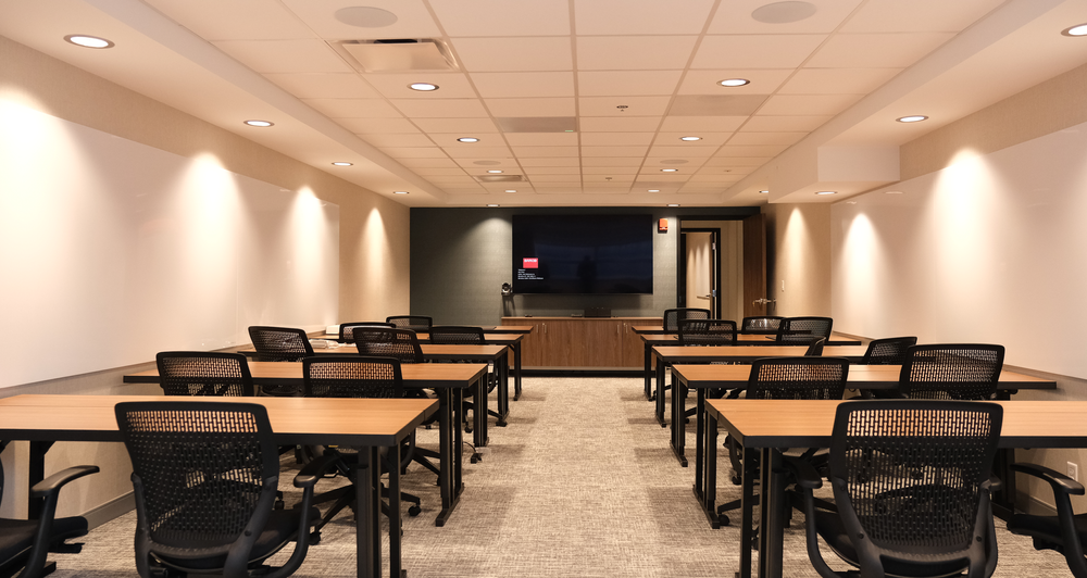 A conference room with 12 desks, 24 black mesh chairs, a large TV on the back wall, and a door to the side.