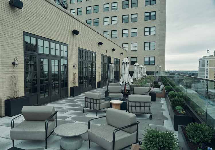 Rooftop patio with chairs, tables, and umbrellas, surrounded by potted plants and a city skyline in the background.