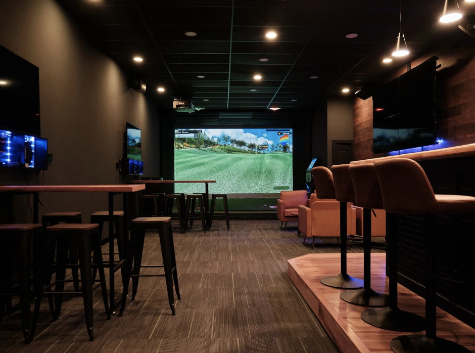 Indoor golf lounge with multiple TVs, some displaying golf courses, seating areas including barstools and armchairs, and a large golf simulation screen at the back.