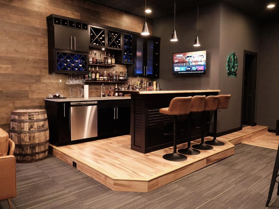 A home bar with a black counter and bar stools, shelves of liquor bottles, wine racks, a small refrigerator, a mounted TV, and decorative lighting.