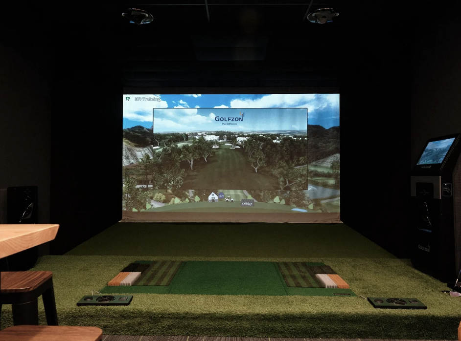 Indoor golf simulator room with a large screen displaying a virtual golf course, artificial grass mat for golf practice, and some seating.