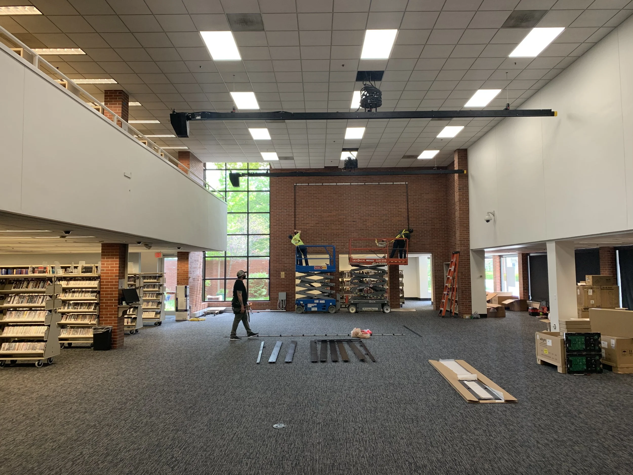 Library interior with workers installing equipment, shelves of books on the left, and a large window at the back letting in natural light.