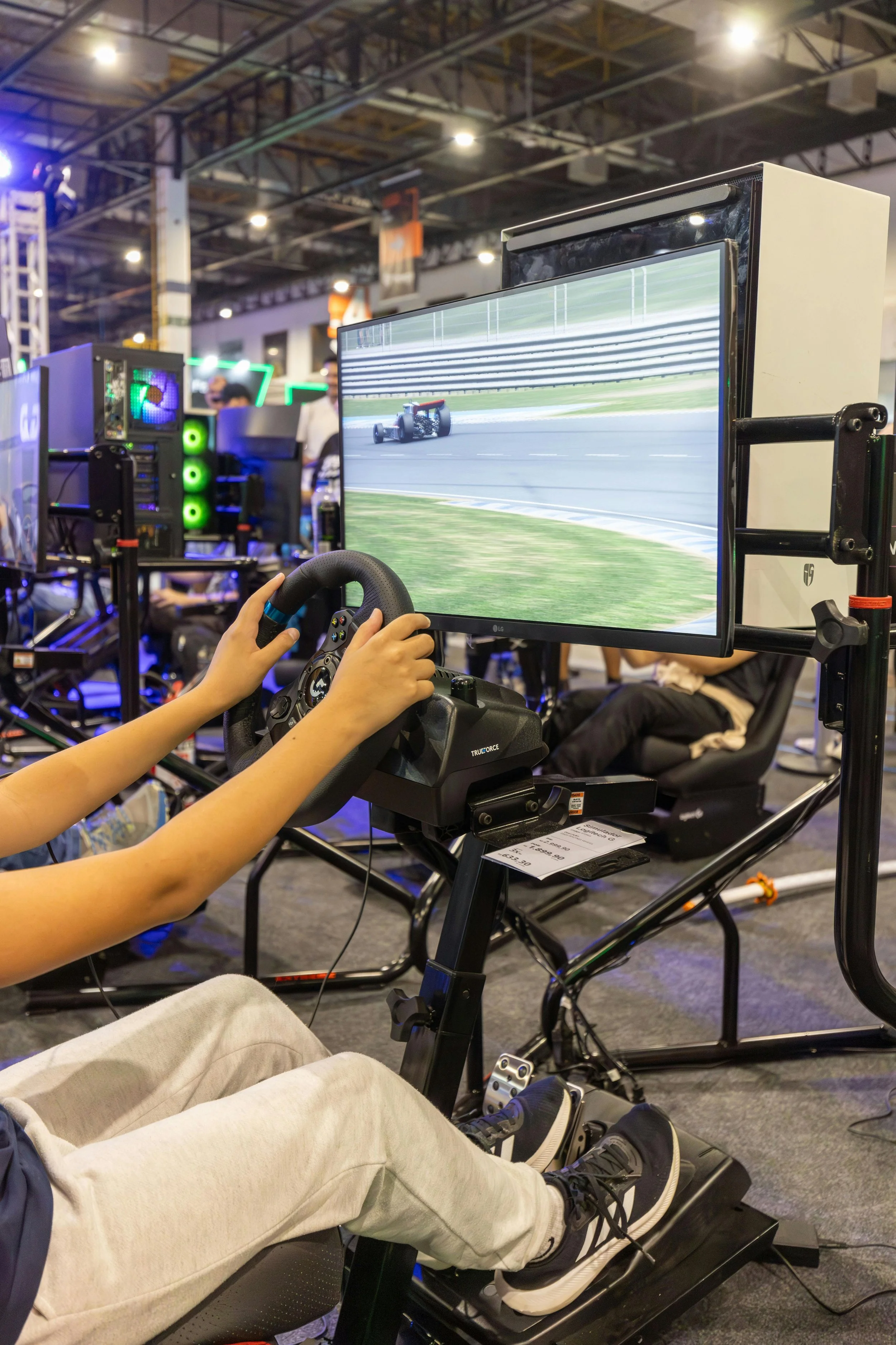 Person playing a racing video game with a steering wheel controller at a gaming event, with a racing car on the screen and gaming PCs around.
