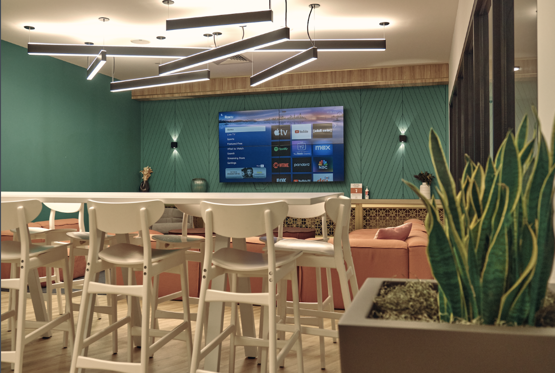 Modern interior lounge area with a wall-mounted TV displaying streaming apps, white high chairs around a bar-height table, peach-colored sofa in the background, and green plants in planters.