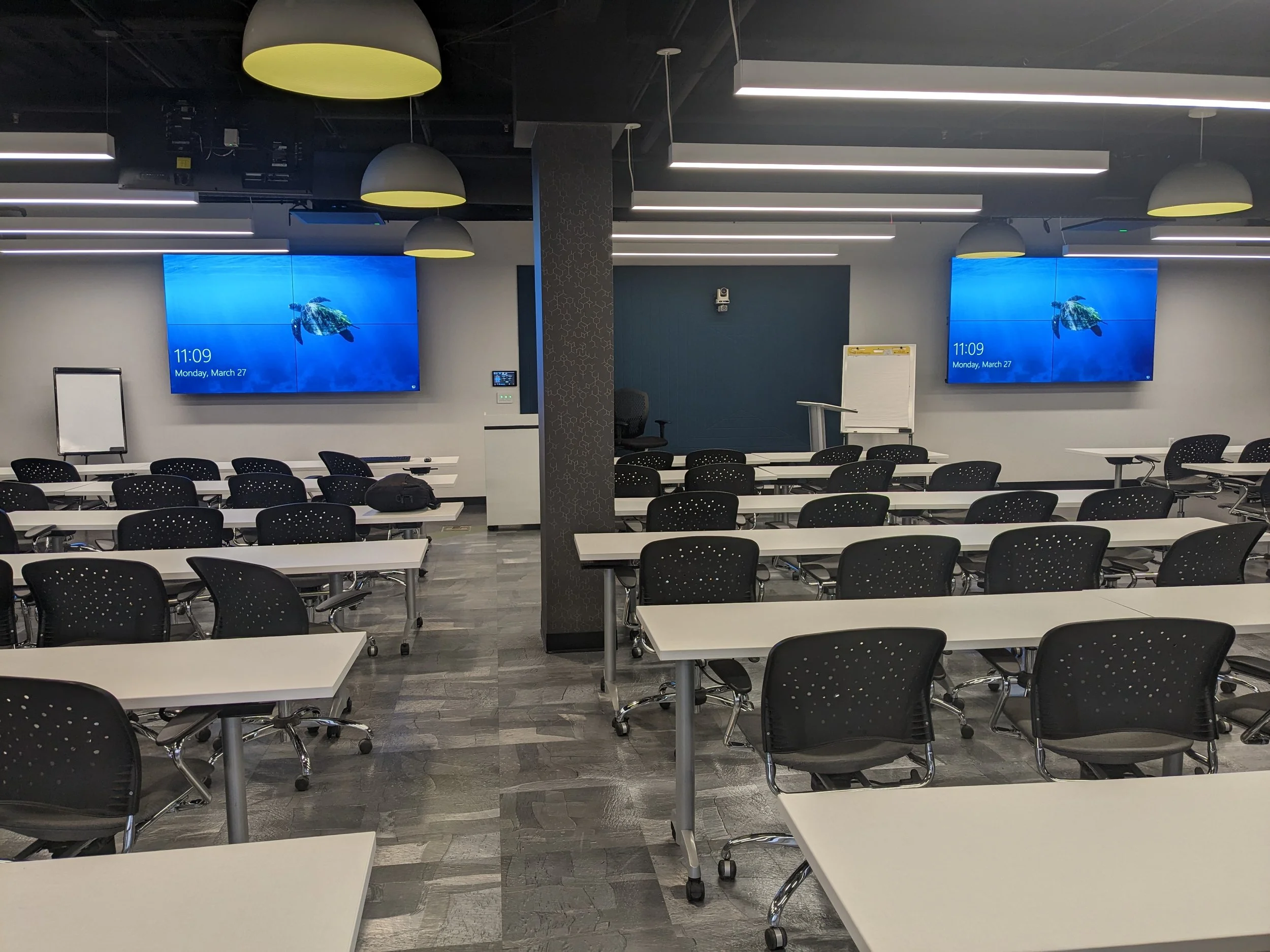 A modern conference room with multiple rows of white tables and black chairs. On the wall, there are two large digital screens showing an underwater scene with a turtle. The room has a gray and black ceiling with hanging lights and a patterned column