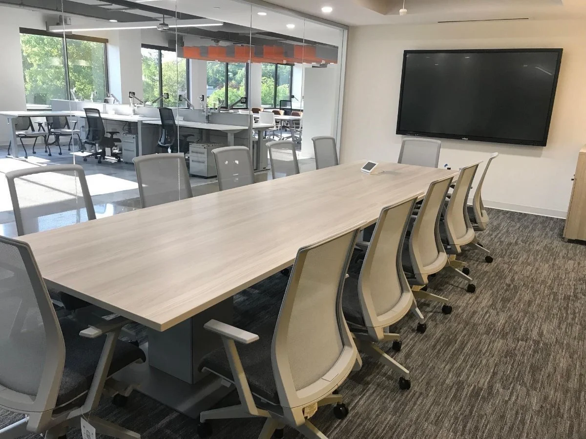 Conference room with a long table and multiple chairs, a large flat-screen television on the wall, and a glass-walled office or meeting space with work desks and chairs visible through the glass.