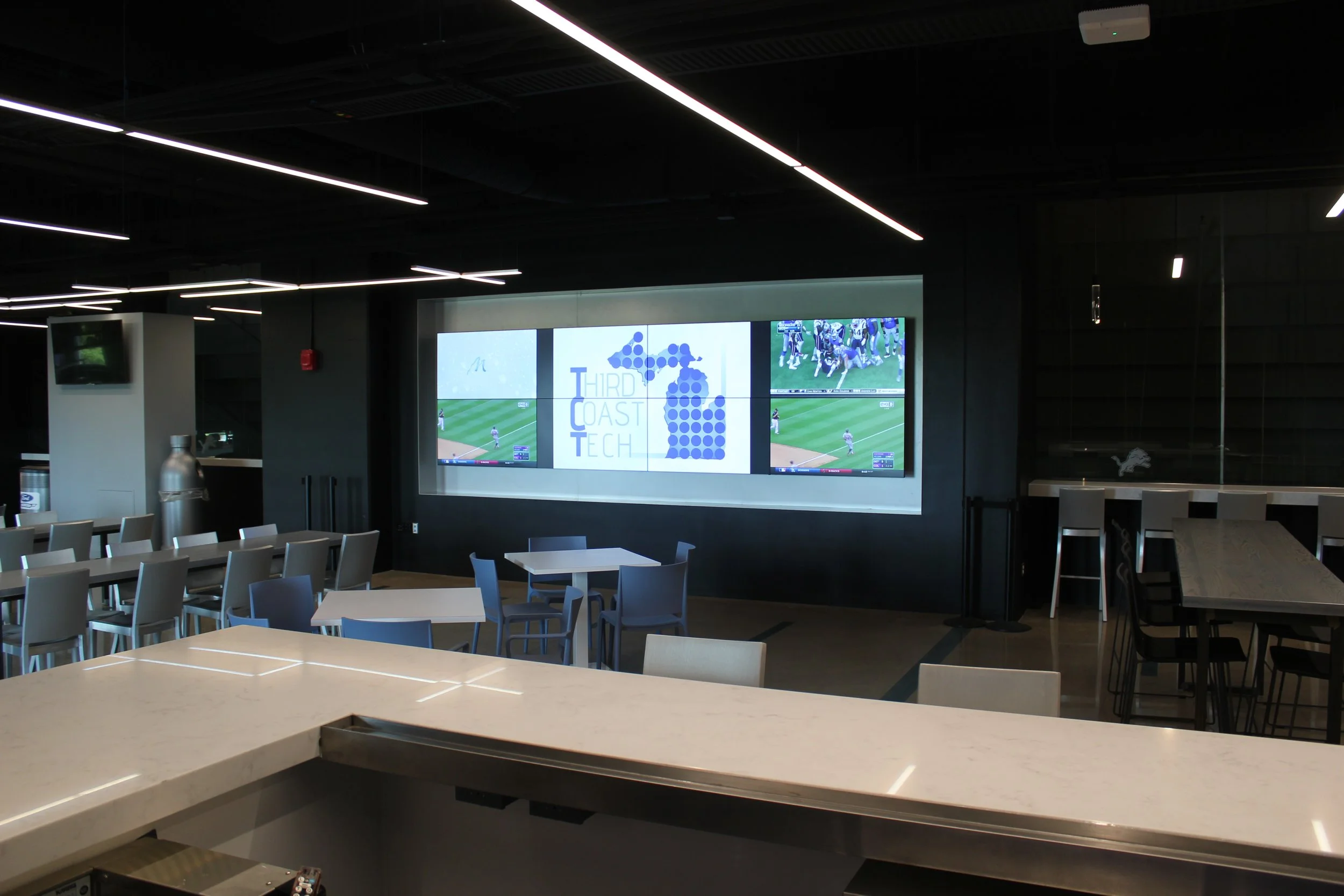 Modern indoor sports lounge with multiple television screens showing a football game, minimalist seating with white chairs and tables, and sleek black ceiling with linear lighting.