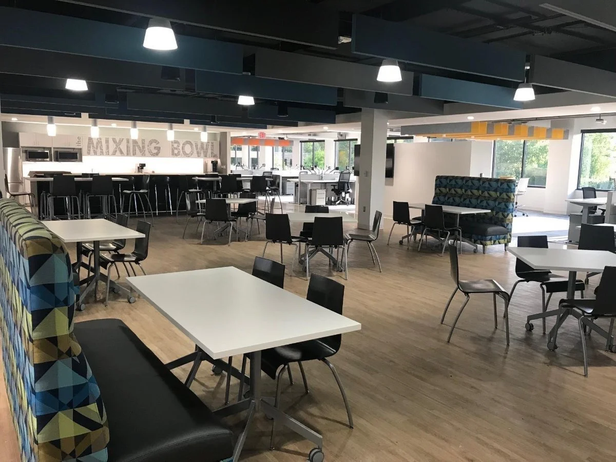 Modern cafeteria with white tables, black chairs, and booths with geometric patterned upholstery, large windows, and sleek lighting.