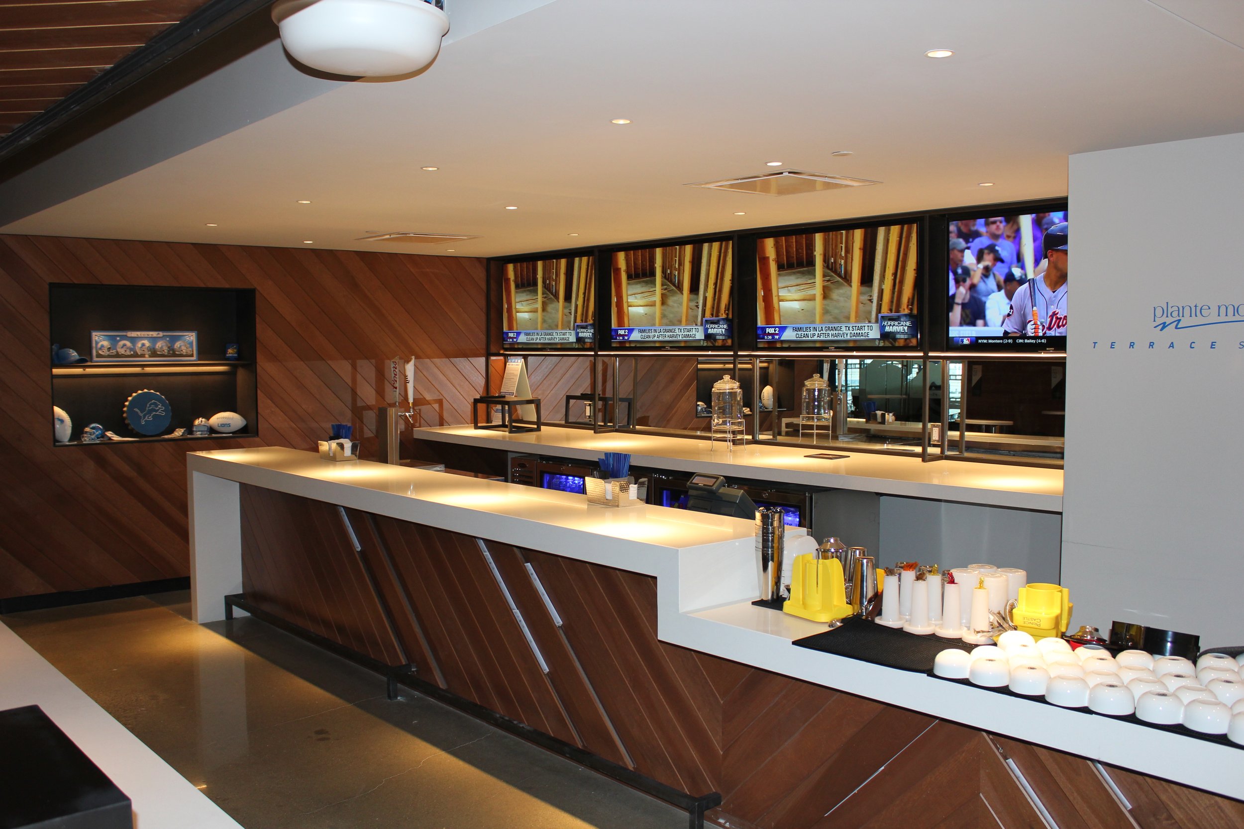 A bar area with a sleek white counter, various condiments and cups, and a wooden wall with multiple television screens showing a baseball game.