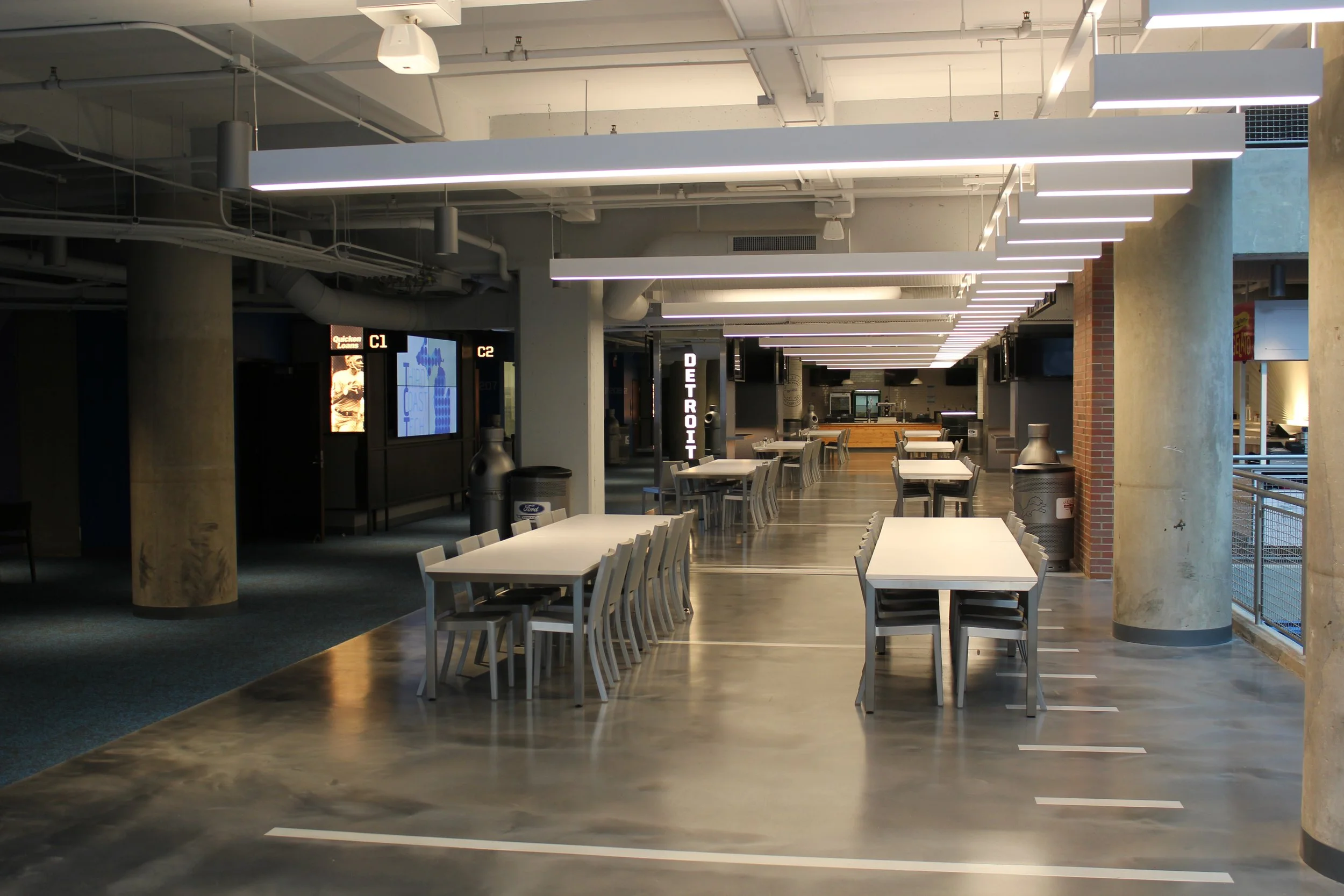 Empty indoor dining area with white tables, white chairs, concrete columns, and modern linear ceiling lighting.