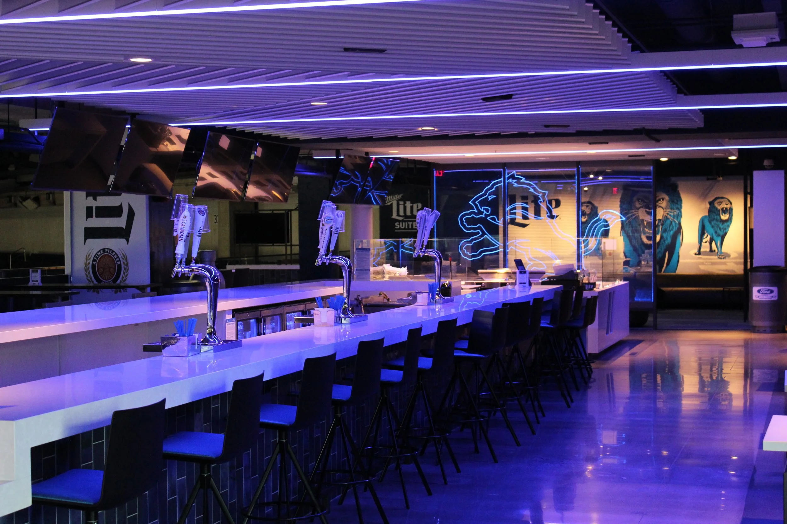 An empty bar or lounge area illuminated by blue lighting, with high white counter tables, black chairs, and several TV screens hanging from the ceiling. Neon lion artwork is visible on the wall behind the bar.