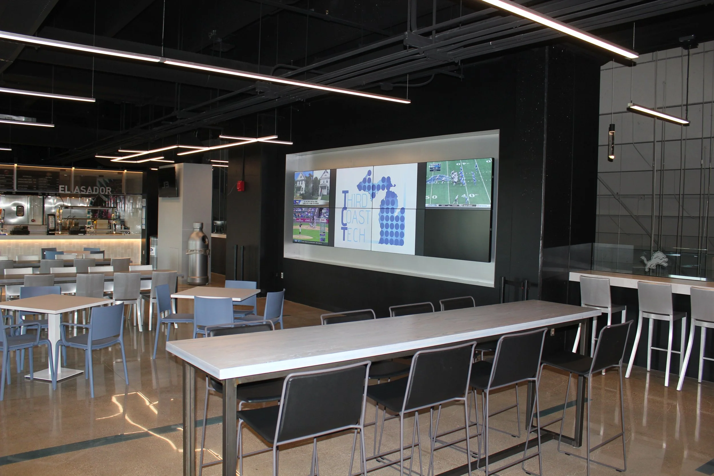 Interior of a modern sports bar or lounge with tables, chairs, large monitors displaying sports games, and contemporary lighting fixtures.