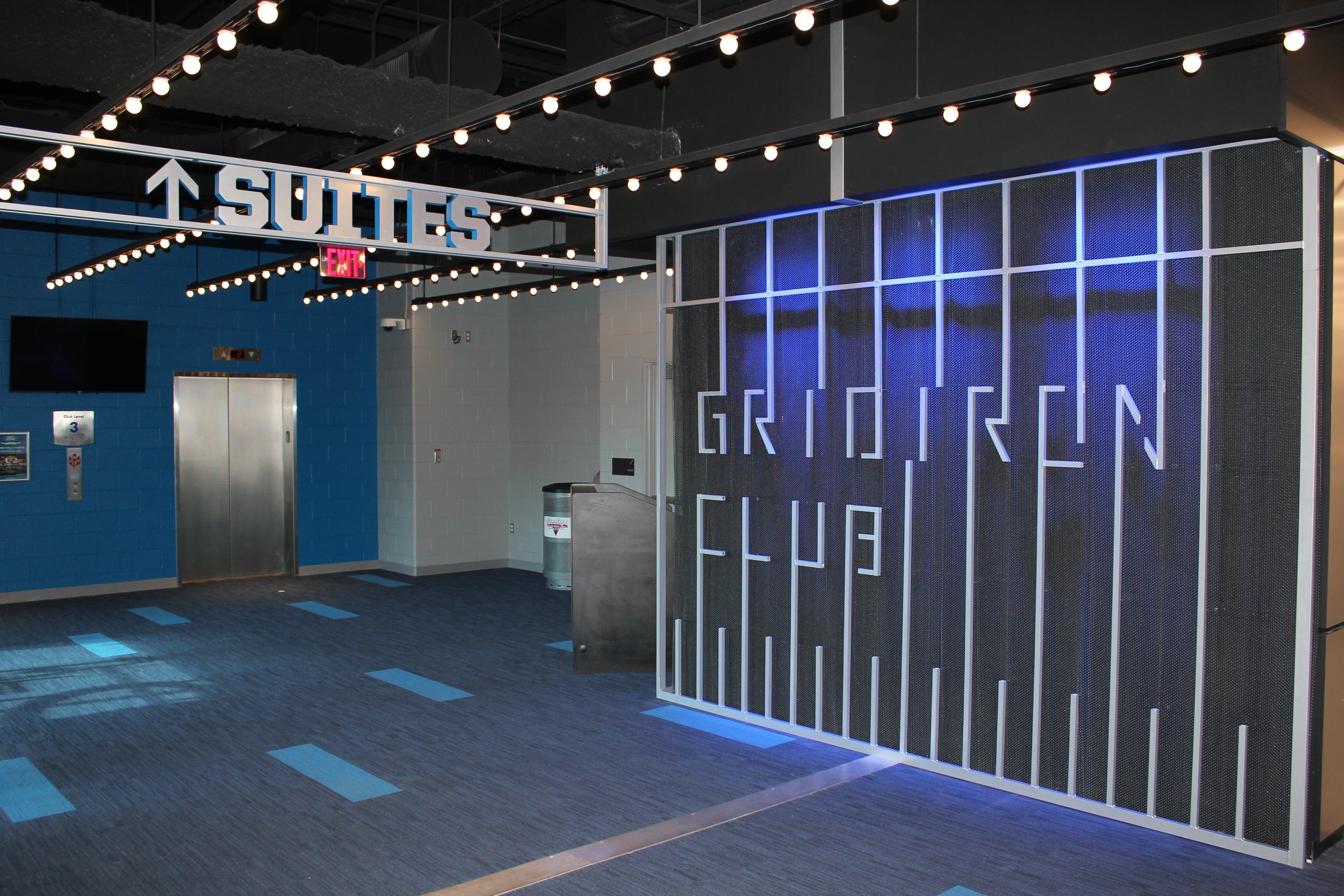 Interior of a modern theater or event space with a sign overhead that says 'SUTIES'. There are light bulbs hanging from the ceiling, a blue brick wall, an elevator door, and a digital sign.