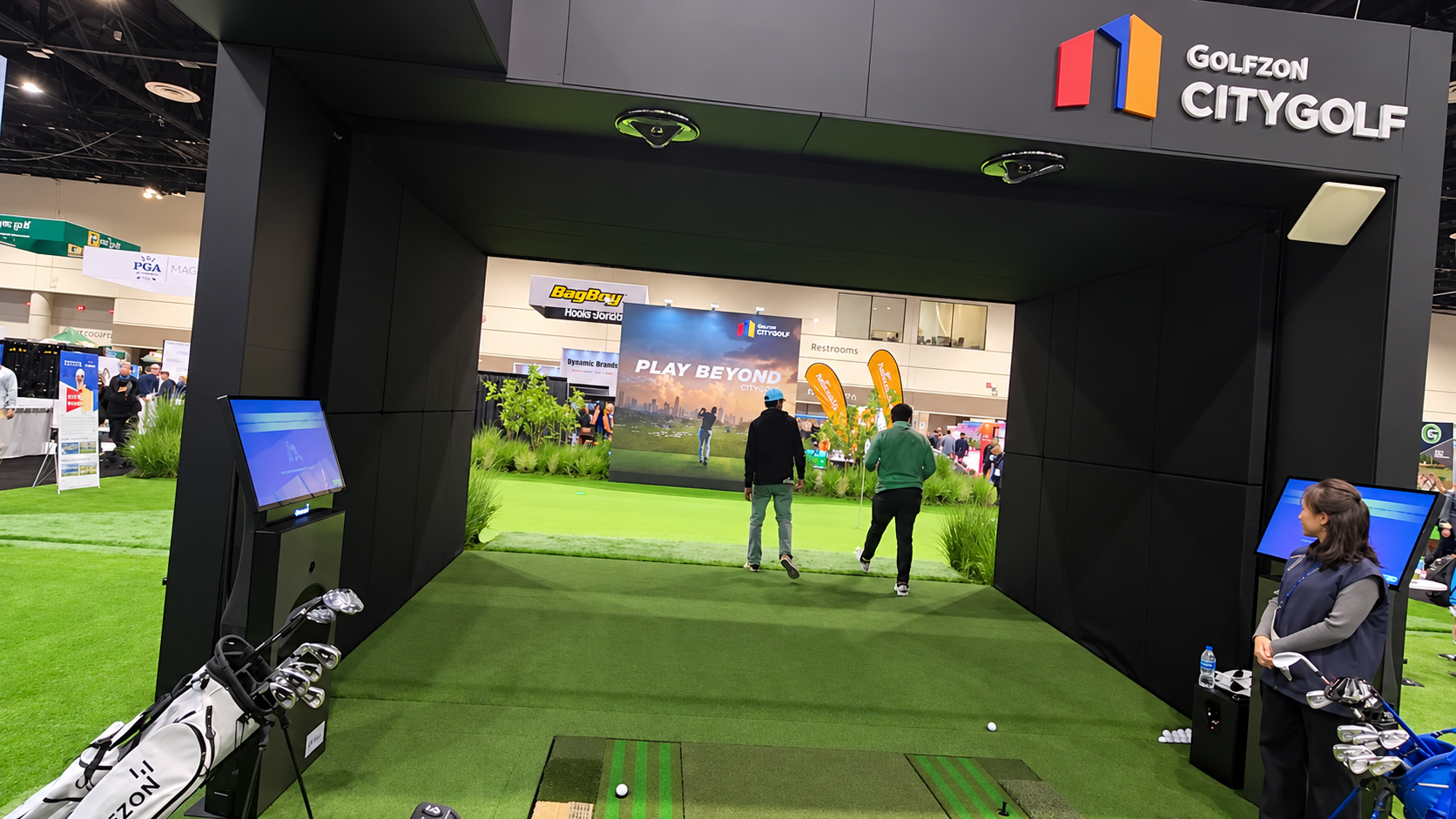 Indoor golf exhibition space with a large black archway entrance labeled "GOLFZON CITYGOLF". Inside, there are area with green artificial grass, golf simulators, and a screen displaying "PLAY BEYOND". A woman is standing near golf clubs, and two men 