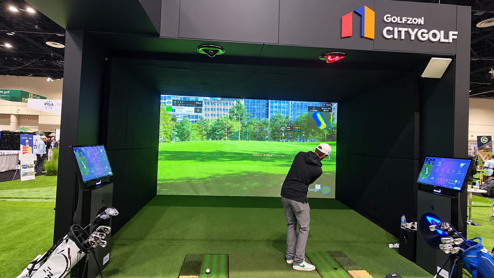 An indoor golf simulator setup at a golf expo with a large digital screen displaying a virtual golf course, a person in a black jacket and white cap practicing their swing, and golf clubs positioned in a golf bag on the left and right sides.