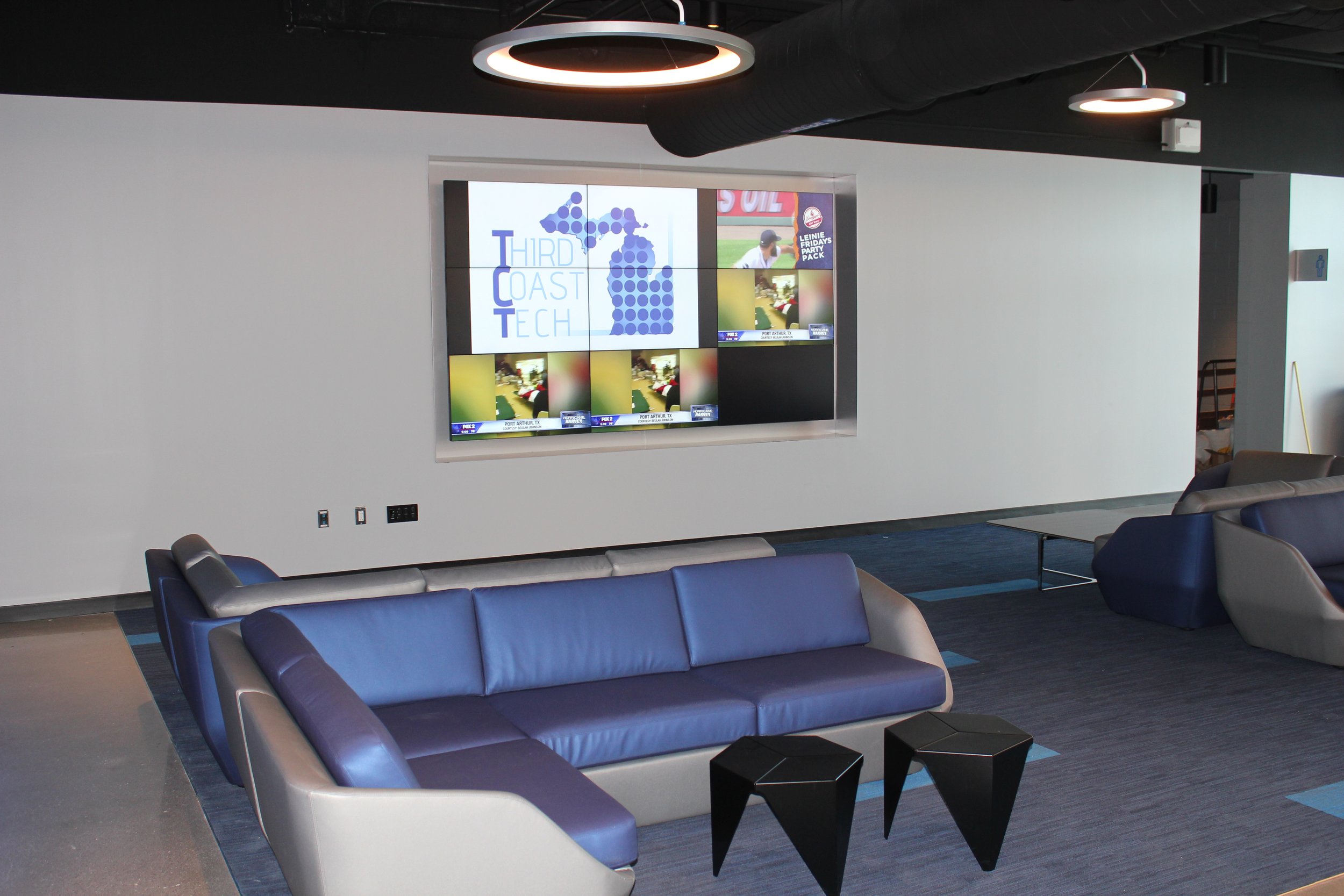 Modern lounge area featuring a color-blocked sofa in blue and gray, two small black tables, and a wall-mounted digital screen displaying various images and graphics. The lounge has dark flooring and a ceiling with black and white circular light fixtu