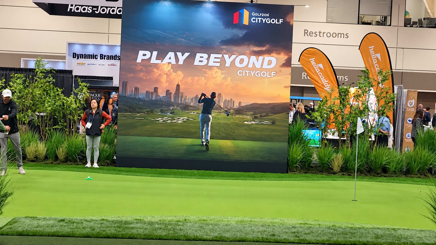 Indoor golf simulation area for a golf tournament or event, featuring a large banner that says 'Play Beyond' with an image of a golfer and city skyline, surrounded by people, flags, and plants.