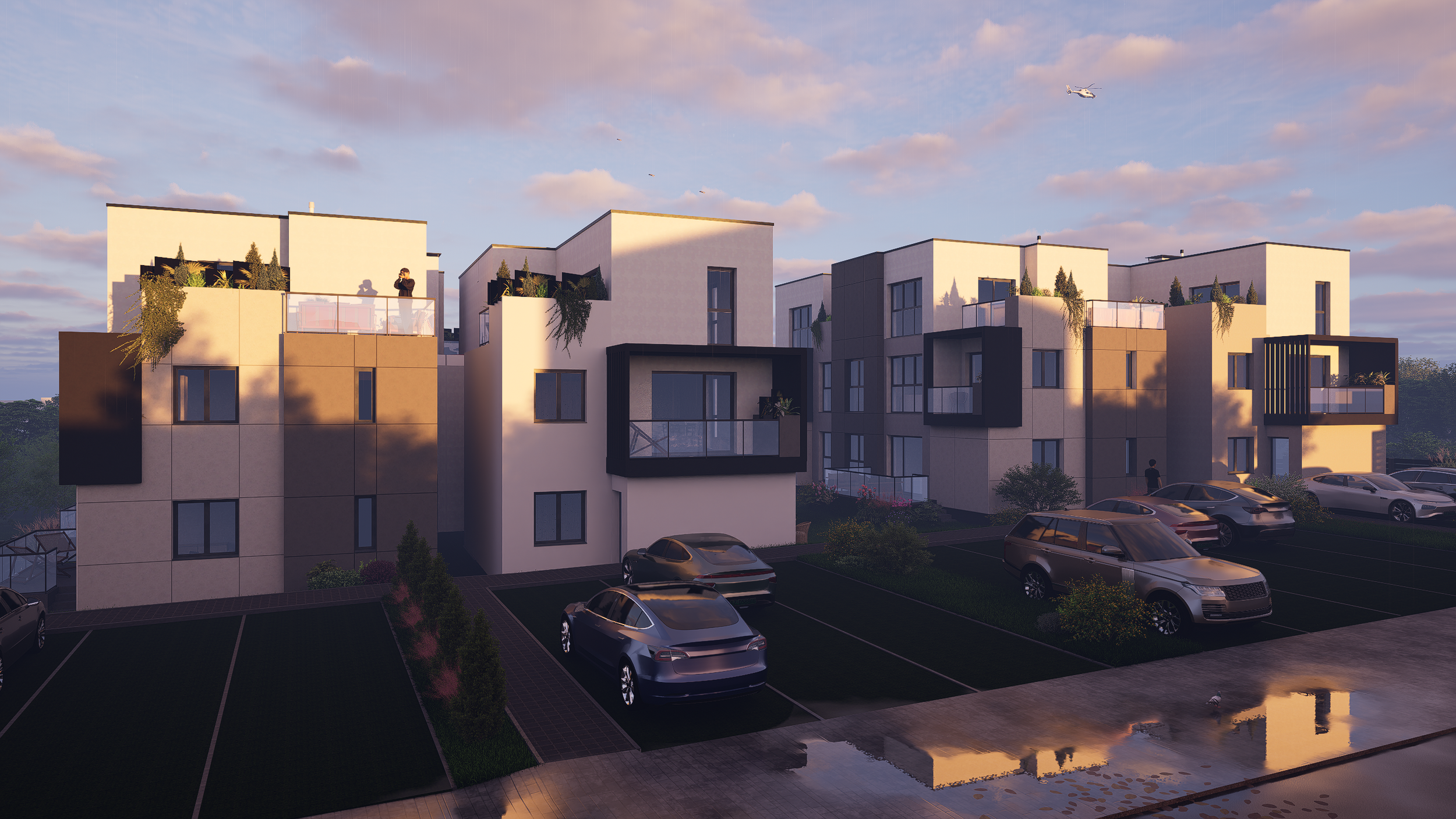 Modern apartment complex with multiple white and beige buildings, parking lot filled with cars, and a partially flooded area reflecting the buildings at sunset. A drone or helicopter is visible in the sky.