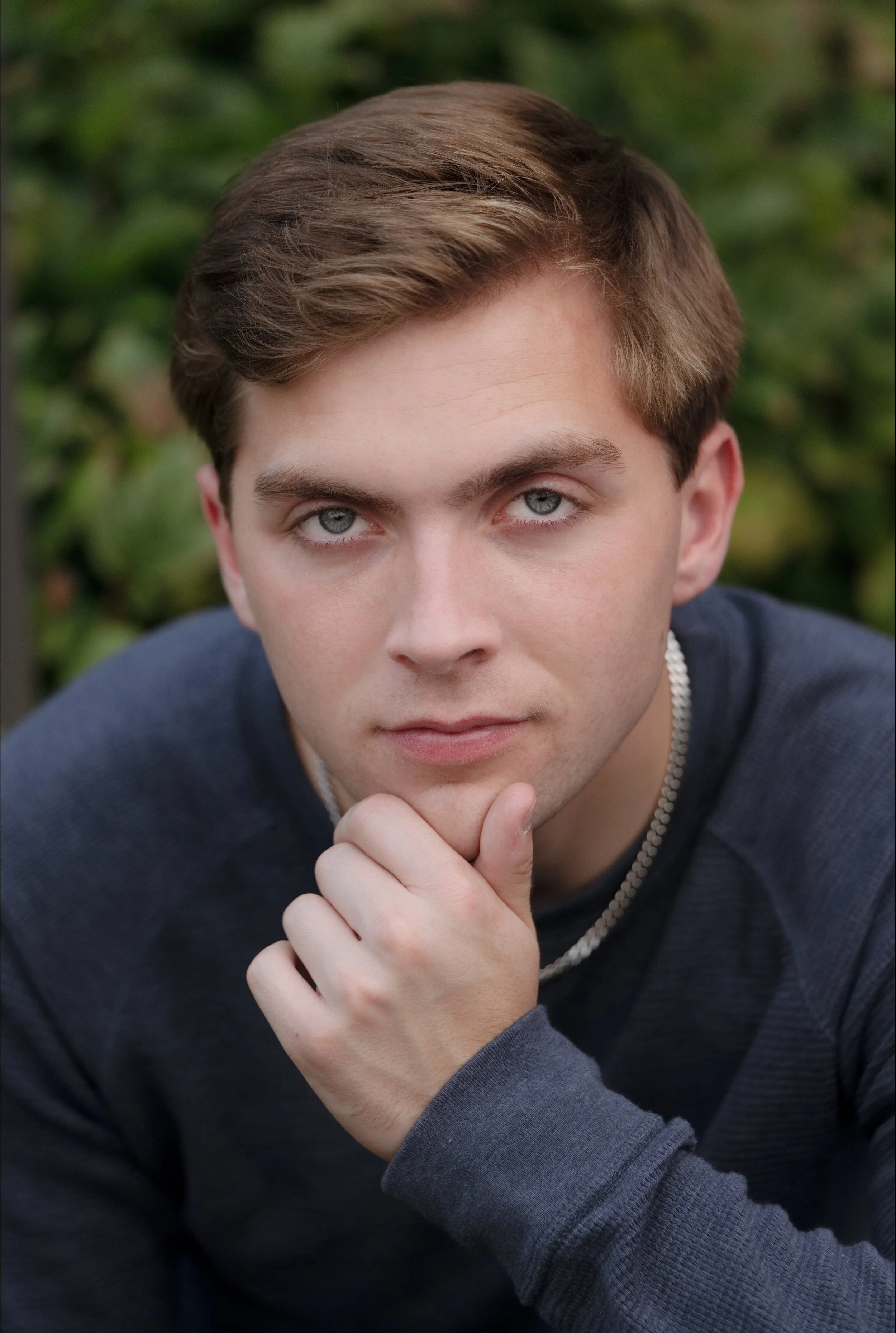 Close-up of a young man with light brown hair and blue eyes, resting his chin on his hand and looking into the camera, outdoors with blurred greenery in the background.