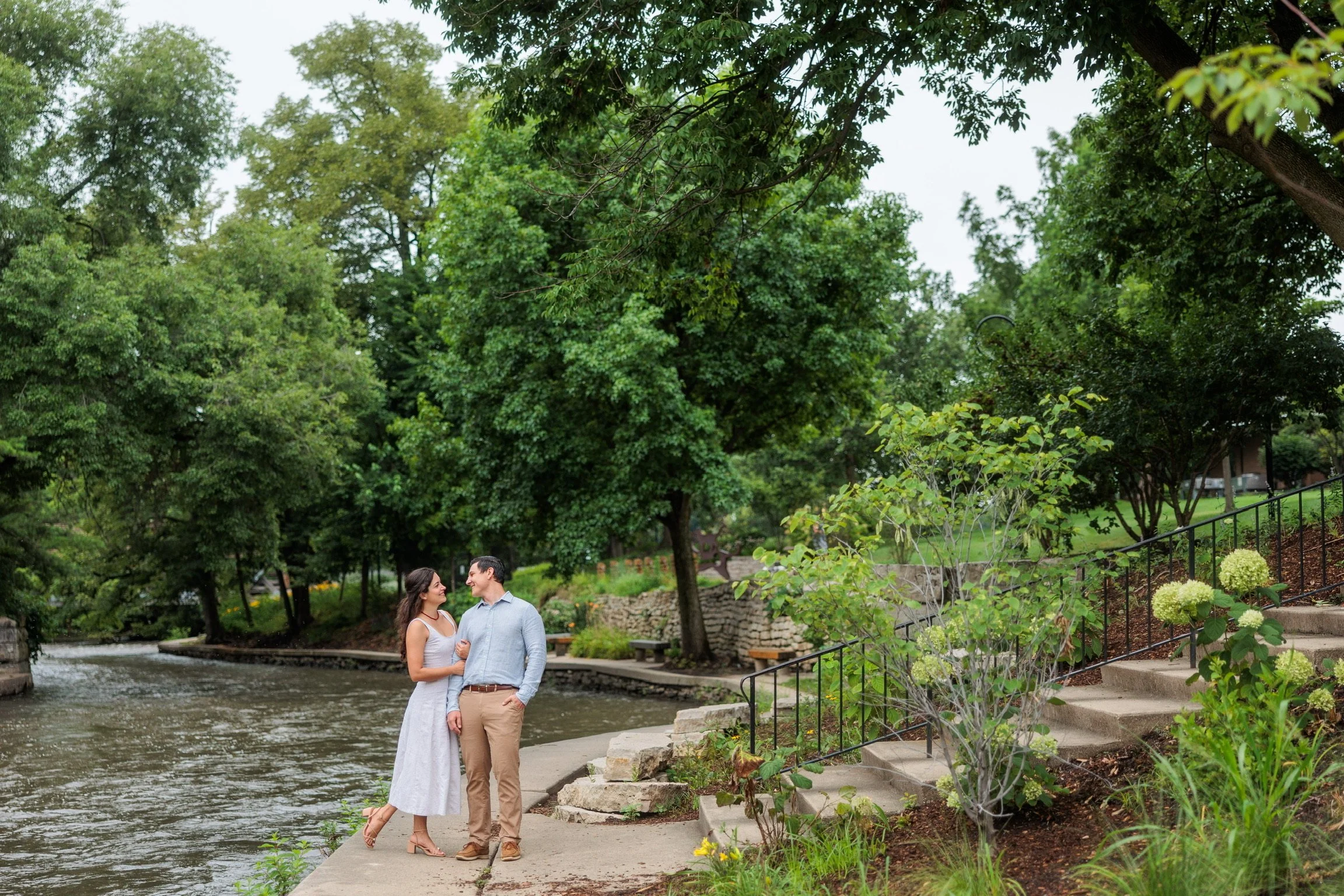 A couple stands together by a river in a park, surrounded by lush green trees and flowering bushes, with stone steps leading down to the water and a black metal fence along the path.