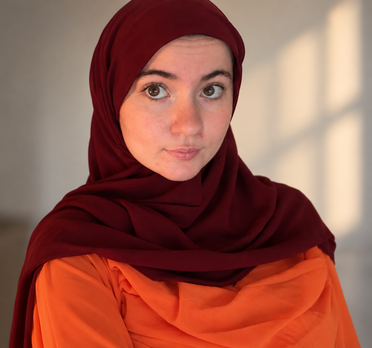 A young woman wearing a red headscarf and orange top, posing indoors with a neutral background.