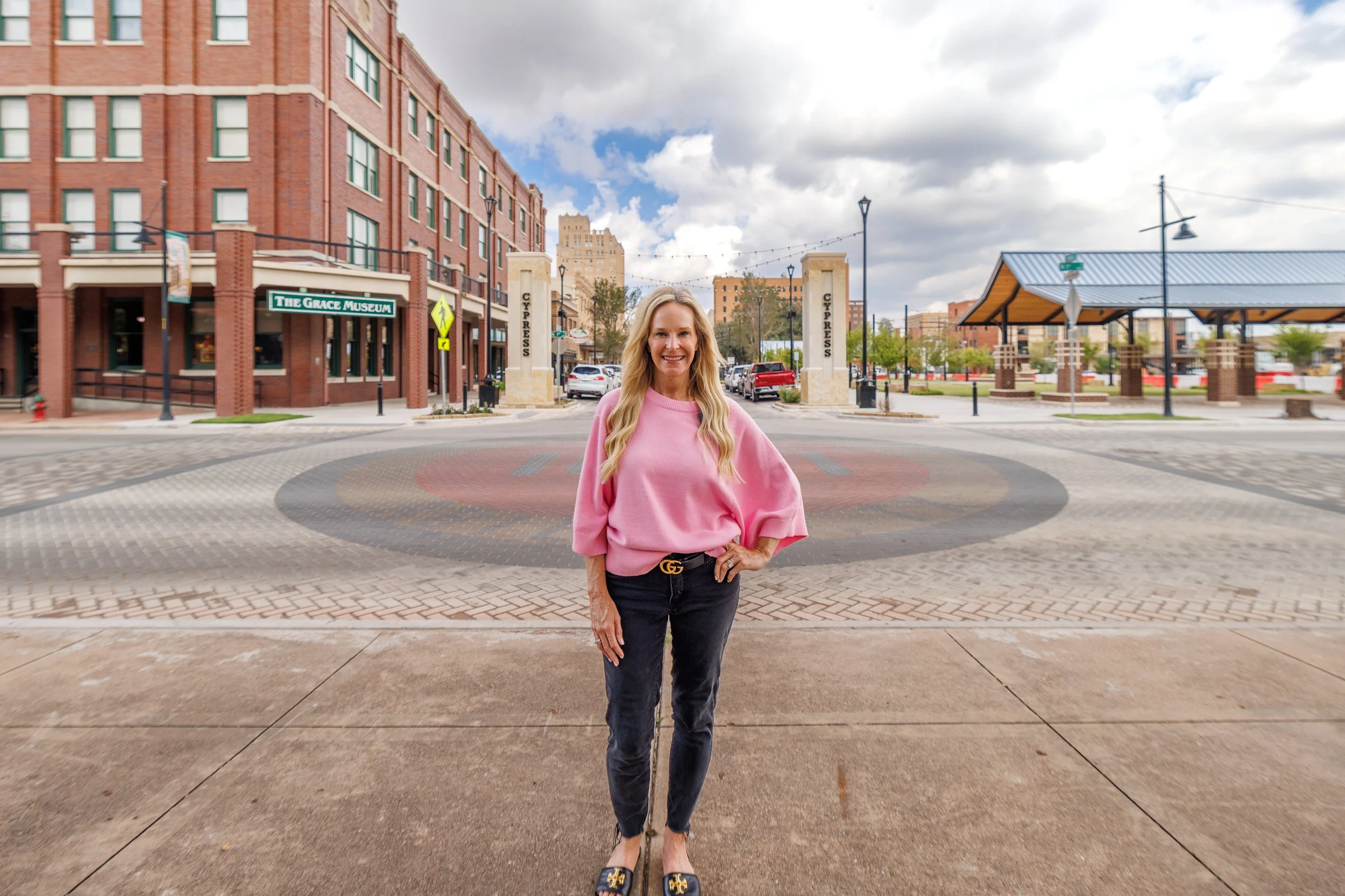 Tonya Harbin, a smiling woman in a pink sweater standing in downtown Abilene, Texas, near The Grace Museum and Cypress Street entrance.