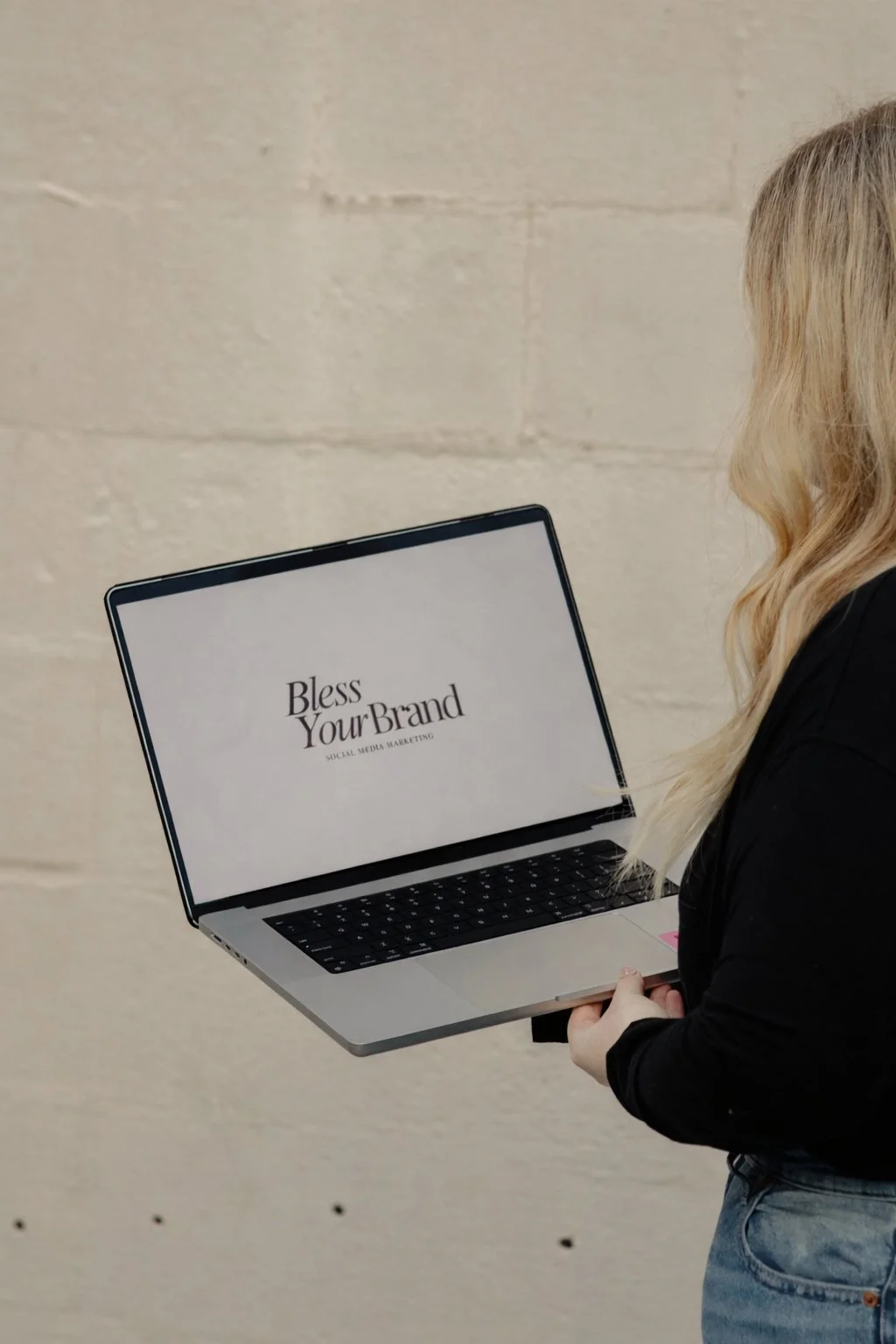 A person with blonde hair is holding a silver laptop with the screen displaying the text 'Bless Your Brand' and 'Social Media Marketing'. The person is standing against a beige brick wall.