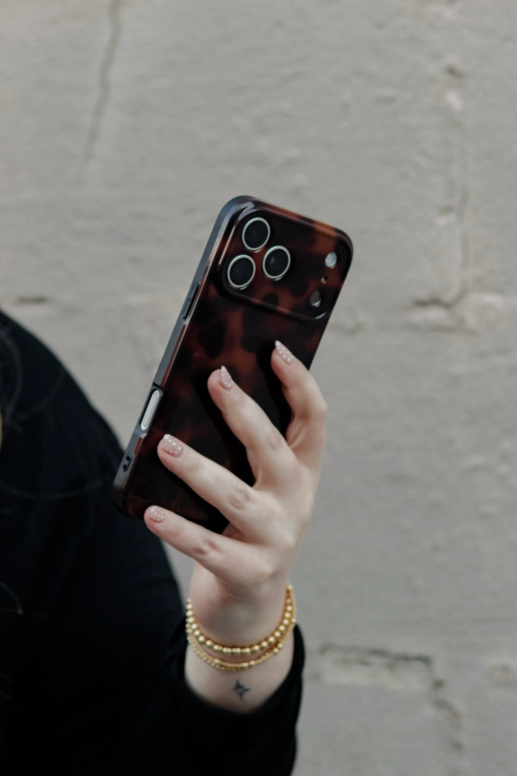Person holding a smartphone with a tortoise shell patterned case, wearing multiple gold bracelets, a star tattoo on wrist, and black clothing, background appears to be a city sidewalk.