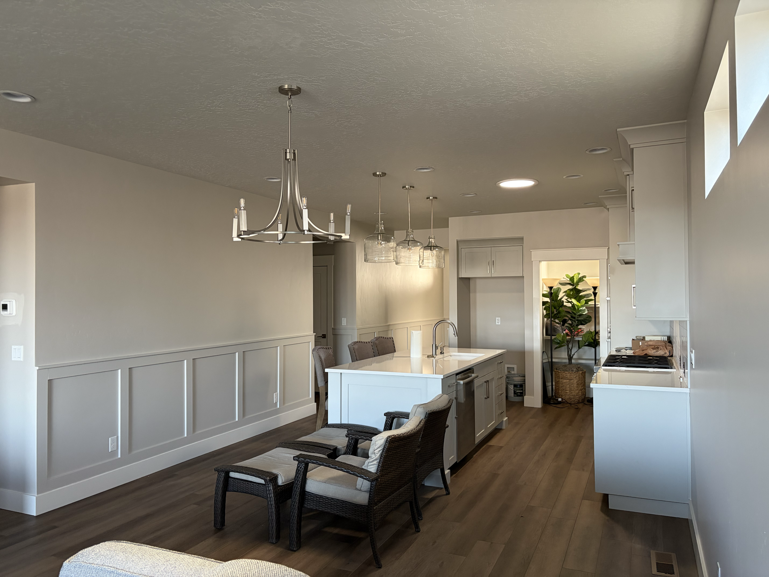 Open plan kitchen and dining area with white kitchen cabinets, island, and modern lighting fixtures, including a chandelier and pendant lights, with wood flooring and some chairs. Residential interior painting in Caldwell, Idaho