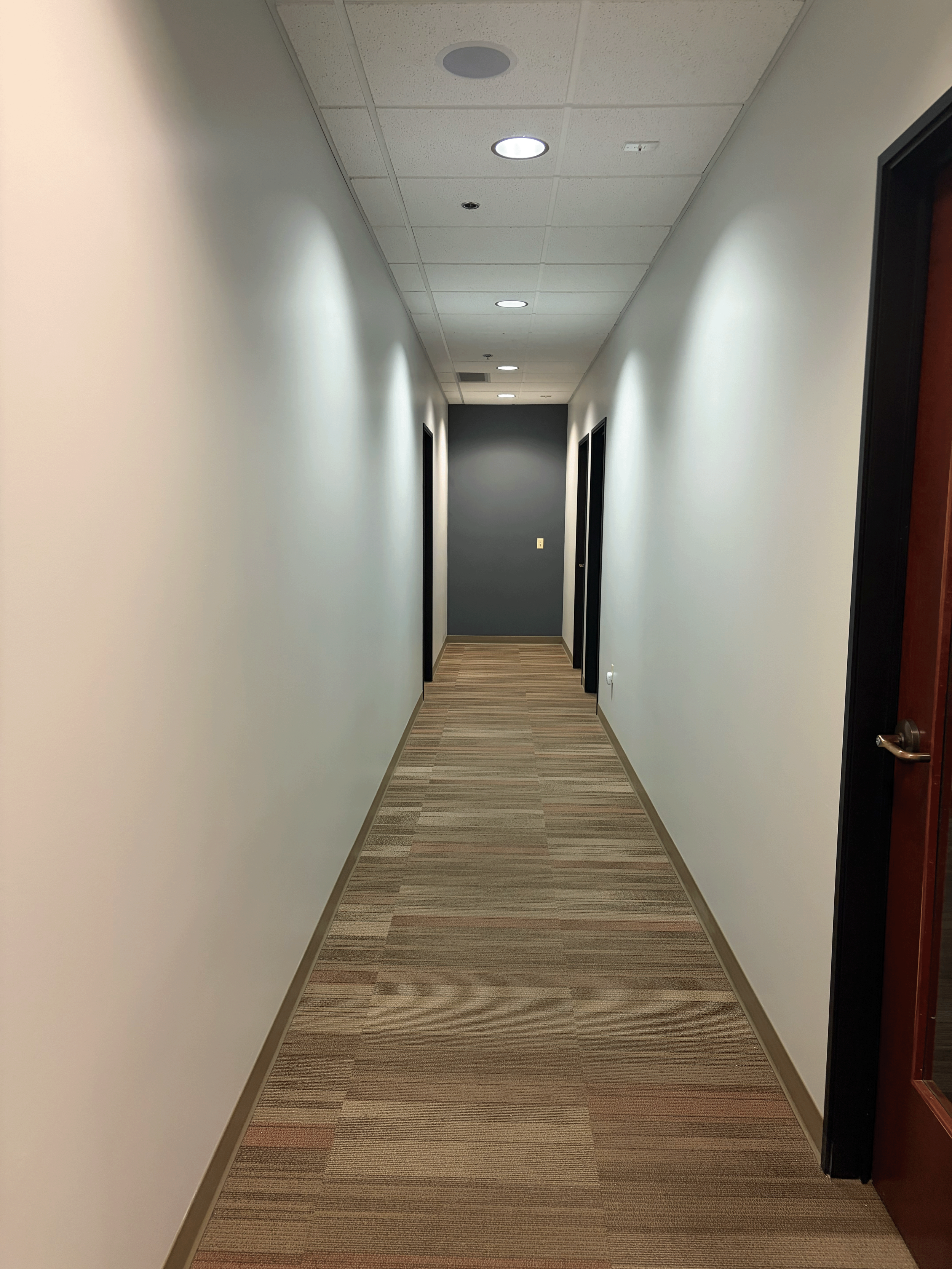 A long, narrow hallway with beige and brown striped carpet, off-white walls, and a gray accent wall at the end. There are three circular ceiling lights along the ceiling. Commercial office interior painting in Boise, Idaho